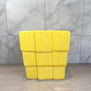 Puede incluir: Una maceta de cerámica amarilla brillante con un diseño texturizado de rayas verticales. La maceta tiene forma rectangular con una apariencia escalonada y apilada. El fondo es una superficie gris claro y una pared de azulejos blancos y grises.
