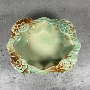 Puede incluir: Un cuenco de cerámica con interior verde claro y borde decorativo con detalles florales marrones y verdes. El cuenco tiene una forma geométrica única y está sobre un fondo gris.