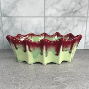 May include: A decorative ceramic bowl with a scalloped rim. The bowl has a light green base with a dark red, dripped glaze around the rim. The rectangular bowl has a wavy edge.