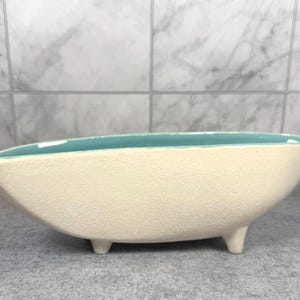 May include: An elongated, boat-shaped ceramic bowl featuring a textured, off-white exterior and a smooth, vibrant turquoise interior. The vessel is supported by two small, sturdy feet at the base, creating a raised profile with a pointed end.