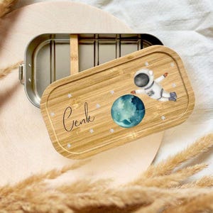 May include: A stainless steel lunchbox with a bamboo lid featuring an astronaut floating near a blue planet. The lid is engraved with the name "Cenk" and small white stars. The lunchbox is open, revealing the interior.