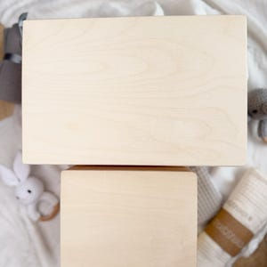 May include: Two unfinished wooden boxes, one large and rectangular, the other smaller and square. The boxes are light-colored with visible wood grain. The boxes are on a white surface with baby toys.