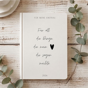 May include: A white hardcover journal with black text that reads F&Uuml;R MEINE EHEFRAU, F&uuml;r all die Dinge, die mein [heart symbol] dir sagen m&ouml;chte, and 2026. The journal has a clean, minimalist design with a smooth finish and centered typography.