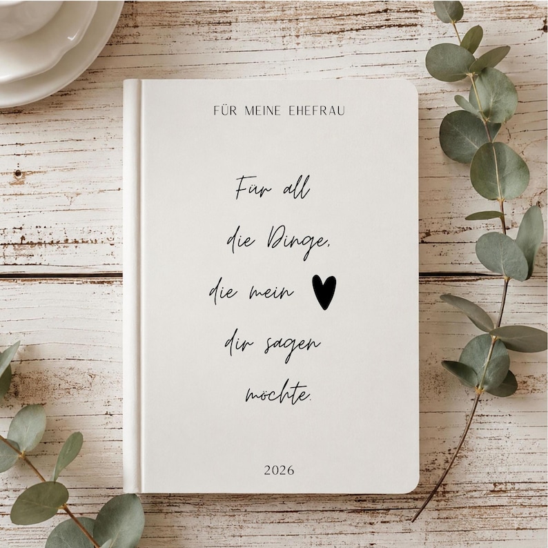 May include: A white hardcover journal with black text that reads F&Uuml;R MEINE EHEFRAU, F&uuml;r all die Dinge, die mein [heart symbol] dir sagen m&ouml;chte, and 2026. The journal has a clean, minimalist design with a smooth finish and centered typography.