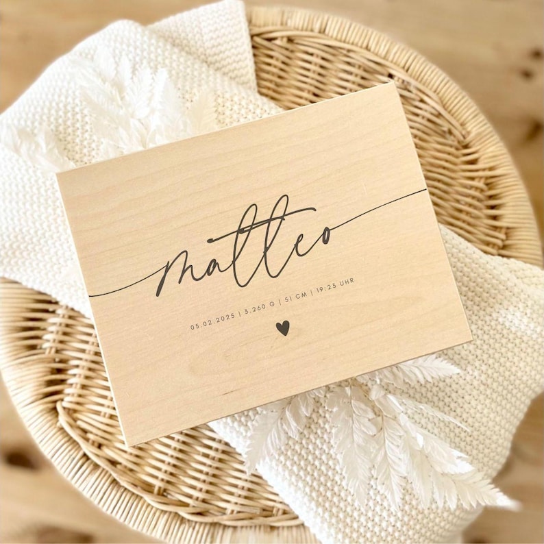 May include: A light-colored wooden plaque with the name "Matteo" in elegant script. The plaque includes the date 05.02.2025, weight 5.260 g, length 51 cm, and time 19:25 Uhr, along with a small heart symbol. The plaque rests on a white blanket.