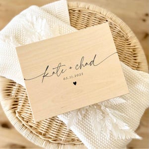 May include: A light-colored wooden box with the names "Kate + Chad" in elegant script, the date "05.11.2025", and a small heart. The box rests on a white blanket and a woven basket.