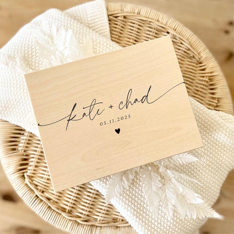 May include: A light-colored wooden box with the names "Kate + Chad" in elegant script, the date "05.11.2025", and a small heart. The box rests on a white blanket and a woven basket.