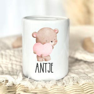 K&ouml;nnte beinhalten: Wei&szlig;e Keramik-Spardose mit einer Aquarell-Illustration eines braunen Teddyb&auml;ren, der ein rosa Herz h&auml;lt. Der Name "ANTJE" ist in schwarzen Blockbuchstaben darunter gedruckt. Die Spardose hat eine zylindrische Form.