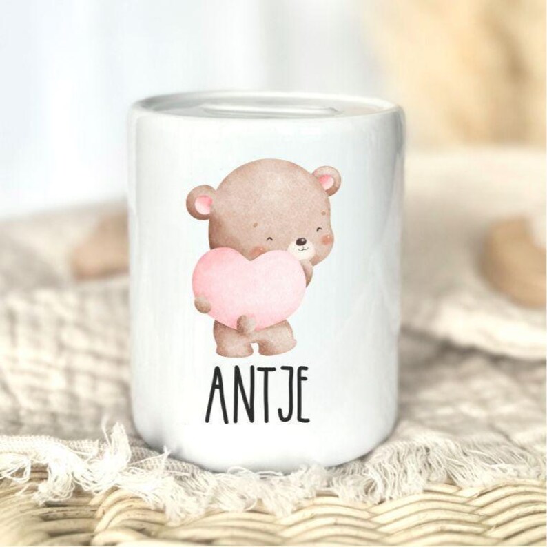 K&ouml;nnte beinhalten: Wei&szlig;e Keramik-Spardose mit einer Aquarell-Illustration eines braunen Teddyb&auml;ren, der ein rosa Herz h&auml;lt. Der Name "ANTJE" ist in schwarzen Blockbuchstaben darunter gedruckt. Die Spardose hat eine zylindrische Form.