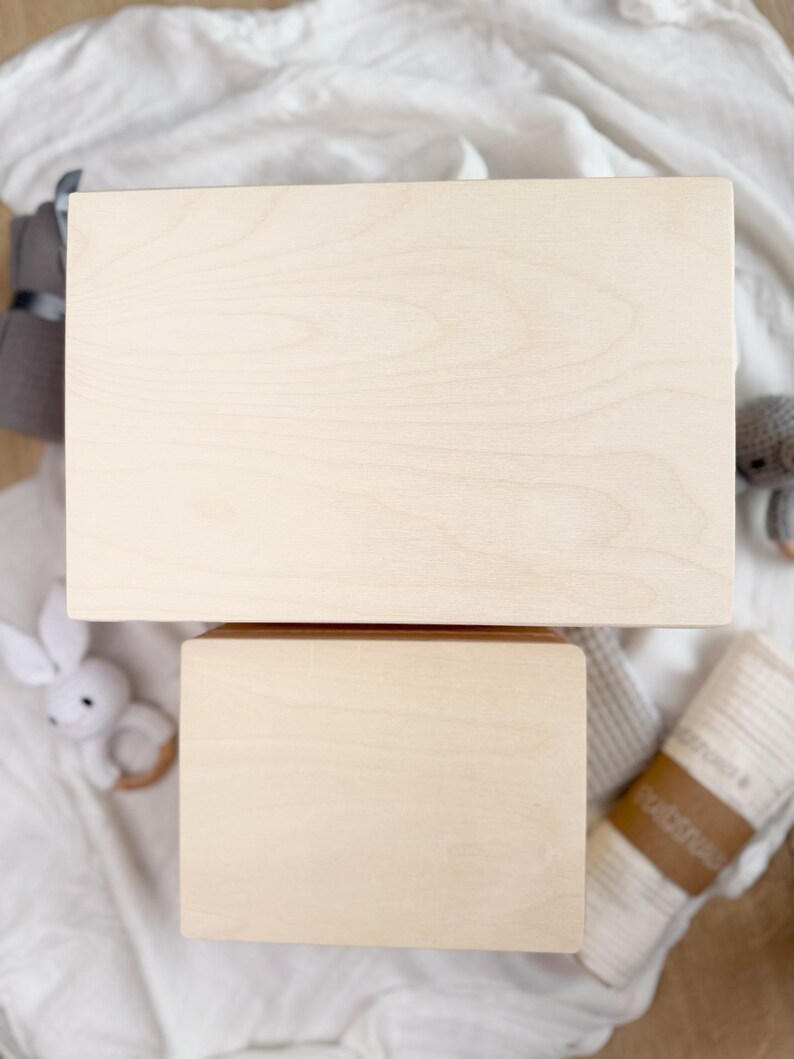 Puede incluir: Dos cajas de madera sin terminar, una rectangular y otra cuadrada, apiladas sobre una superficie blanca. Las cajas tienen un veteado de madera claro. Se ven un juguete de conejo blanco y un peluche gris.