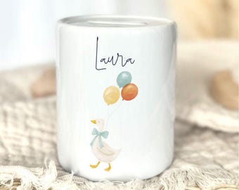 Könnte beinhalten: Weiße Keramik-Spardose mit einer Illustration einer Gans, die Luftballons hält. Der Name "Laura" ist in handschriftlicher Schrift auf der Vorderseite geschrieben. Die Ballons sind gelb, orange und türkis.