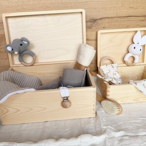 May include: Two open wooden boxes filled with baby items. One box contains a gray blanket, gray fabric, and a pacifier clip. The other box has a white and gray bunny teether, a brush, and a floral print cloth. A gray elephant teether is in the first box.