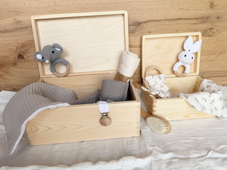 May include: Two open wooden boxes filled with baby items. One box contains a gray blanket, gray fabric, and a pacifier clip. The other box has a white and gray bunny teether, a brush, and a floral print cloth. A gray elephant teether is in the first box.