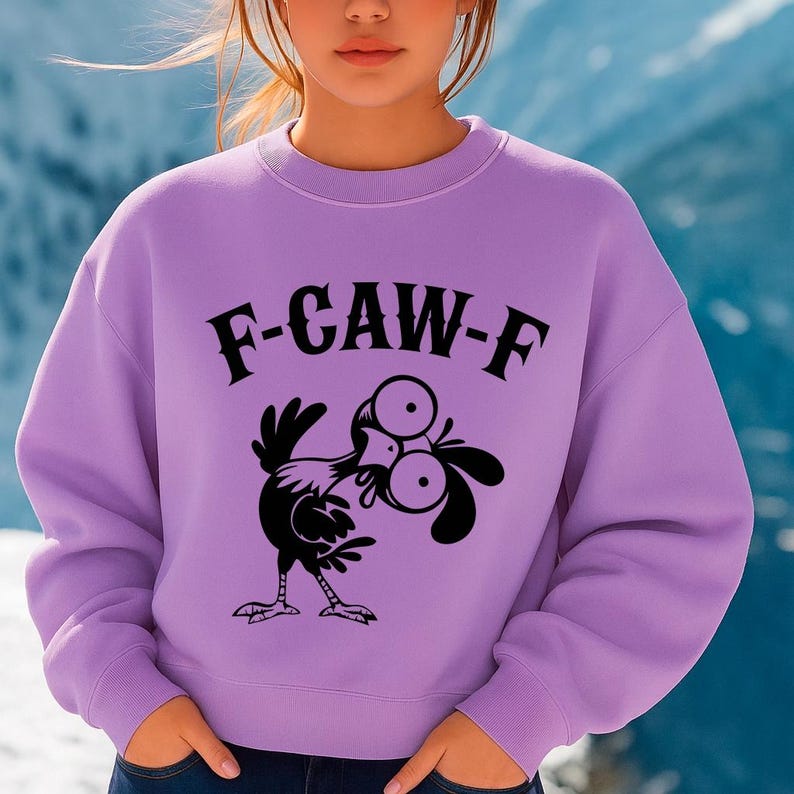 May include: A lilac-colored sweatshirt with the text "F-CAW-F" in black, above a cartoon chicken illustration. The chicken has large eyes and is in a comical pose. The sweatshirt has long sleeves and a crew neck.