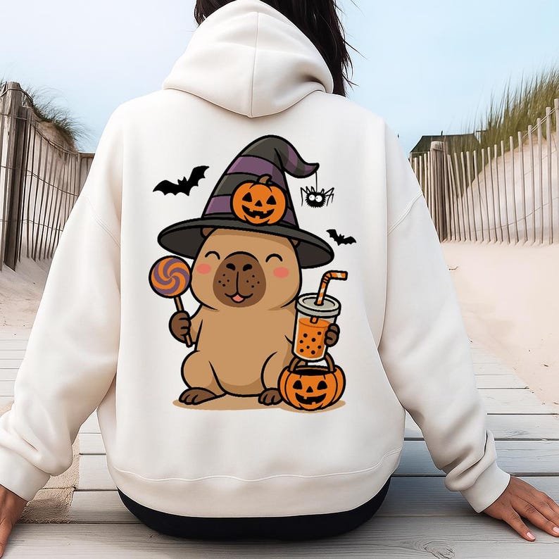 May include: Cream-colored hoodie featuring a Halloween-themed capybara illustration. The capybara wears a witch hat with a pumpkin, and holds a lollipop, a drink, and a jack-o'-lantern. Black bats and a spider complete the design.