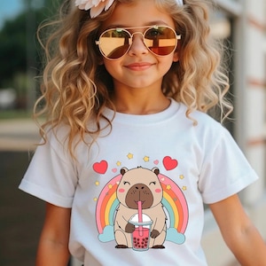 May include: A young person wearing a white t-shirt featuring a cartoon capybara drinking boba tea in front of a rainbow. The shirt has a cute design with hearts and stars. The person is also wearing sunglasses and a flower in their hair.
