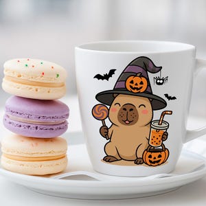 May include: A white ceramic mug featuring a cartoon capybara wearing a witch hat and holding a lollipop and a drink. The mug is on a white saucer with a stack of three macarons in pastel colors. Halloween themed.