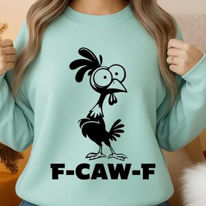 May include: A light teal sweatshirt featuring a black cartoon chicken with large eyes and a comb. The text "F-CAW-F" is printed in bold, black letters below the chicken. The sweatshirt has long sleeves.