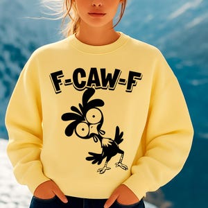 May include: A light yellow sweatshirt with the text "F-CAW-F" in black block letters above a cartoon chicken graphic. The chicken is black and white with large eyes and a surprised expression. The sweatshirt has long sleeves.