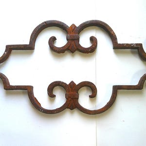 May include: Ornate, rusty brown metal decorative piece with a symmetrical design. Features scrolled ends and fleur-de-lis accents, perfect for adding a vintage touch to decor. The piece is approximately 12 inches wide.