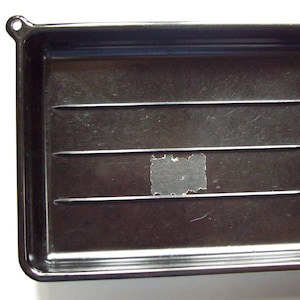 May include: A dark brown rectangular plastic sorting tray featuring three horizontal raised ridges across the center and a small circular hole at the top left corner. A small rectangular adhesive residue patch is visible in the center.