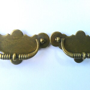 May include: Two antique brass drawer pulls with a decorative design. Each pull features a curved handle with a ribbed texture and a decorative backplate. The pulls are a warm, aged brass color.