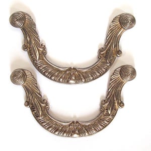 May include: Two ornate, silver-toned drawer pulls with a curved, U-shaped design. Each pull features a scrolled end and a central decorative element. The pulls have a detailed, textured surface and are set against a white background.
