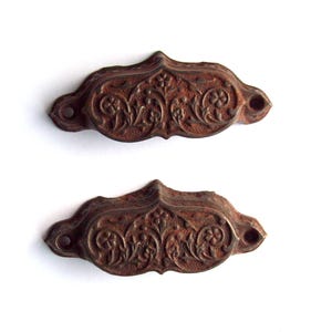 May include: Two antique, ornate drawer pulls with a distressed, rusty brown finish. Each pull features a detailed floral and scrollwork design, with mounting holes on either end. The pulls are set against a white background.