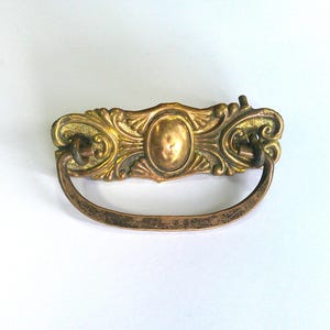 May include: Ornate antique brass drawer pull with a curved handle. The decorative hardware features an oval center and scrolling details. The aged finish gives it a vintage look, perfect for restoring furniture.