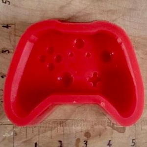 May include: A vibrant red silicone mould shaped like a video game controller. The mould shows the controller's buttons and directional pad. The mould is approximately 10 cm wide and 6.5 cm tall.