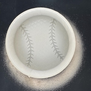 May include: A white baseball-themed coaster with a detailed baseball stitch design. The coaster is round and has a raised edge. The design is a light gray color, and the coaster is set against a dark background.