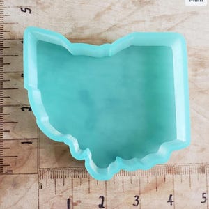 Könnte beinhalten: Eine hellblaue Silikonform in Form des Bundesstaates Ohio. Die Form ist etwa 10 cm breit und 10 cm lang. Die Form liegt auf einer Holzoberfläche mit einem Lineal.