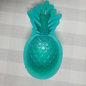 Puede incluir: Un molde en forma de piña de color turquesa. El molde tiene una superficie texturizada con un diseño detallado, incluyendo la corona y el cuerpo segmentado de la piña. El molde está hecho de un material flexible.