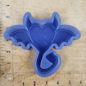 May include: A blue plastic cookie cutter in the shape of a heart with wings and a tail. The cookie cutter is on a wooden surface with a ruler.