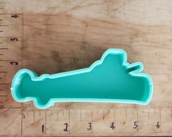 May include: A teal-colored airplane-shaped cookie cutter. The cutter is made of plastic and is approximately 5 inches long. The cutter is on a wooden surface with a ruler.