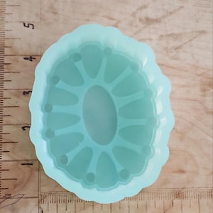 以下が含まれることがあります： 楕円形の中央と放射状の線を持つ、水色のシリコンモールド。 モールドはスカラップエッジで、センチメートル単位の目盛りが付いた木製の表面にあります。