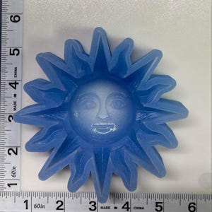 Op de afbeelding: Een blauwe zonvormige mal met een lachend gezicht in het midden. De zon heeft puntige stralen die naar buiten uitsteken. De mal is gemaakt van een doorschijnend materiaal. De afbeelding is van bovenaf genomen.