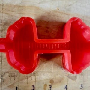 Peut inclure: Un objet rouge vif en forme d'haltère, probablement un moule ou un jouet, est centré dans le cadre. L'objet est fait d'un matériau solide et lisse. Il est posé sur une surface en bois avec une règle pour l'échelle. La forme de l'haltère est clairement définie.