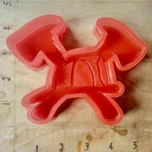 May include: A bright red plastic cookie cutter in the shape of a crab. The cutter is placed on a wooden surface next to a ruler with centimetre measurements.