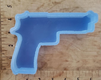 Puede incluir: Un molde de silicona azul claro con forma de pistola. El molde está sobre una superficie de madera con marcas de medición en centímetros. El molde es un contorno detallado de una pistola.