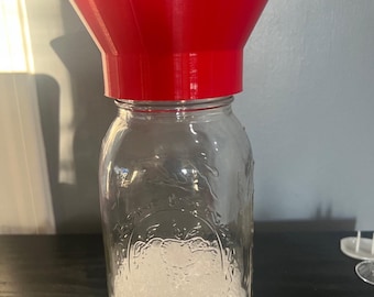 Puede incluir: Un tarro de cristal transparente lleno de pequeñas cuentas blancas, coronado por un embudo rojo brillante. El tarro tiene diseños florales en relieve y está sobre una superficie oscura. El embudo es de plástico.