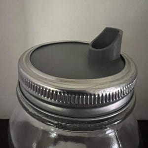 May include: A clear glass jar with a silver metal lid and a gray plastic pour spout. The lid is secured with a threaded metal band. The jar is designed for food storage and dispensing.