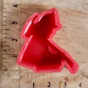 May include: A red plastic cookie cutter shaped like a firefighter with an axe. The cutter is approximately 7.6 cm tall and rests on a wooden surface with a ruler.