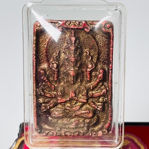 Puede incluir: Un colgante rectangular encerrado en plástico transparente. El colgante presenta una figura detallada de múltiples brazos en tonos marrones y rojos. Tiene un lazo plateado en la parte superior para colgar.