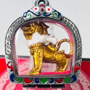 Könnte beinhalten: Eine goldfarbene Löwenfigur mit einem weißen Faden um den Hals, eingefasst in einen silberfarbenen Metallrahmen mit roten, blauen und grünen Emaille-Akzenten. Der Anhänger hat eine klare Schutzhülle und eine Öse.