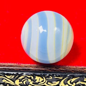 Può includere: Una biglia di vetro sferica caratterizzata da strisce verticali alternate di colore azzurro, bianco e sottili linee gialle. La superficie della biglia è liscia e riflettente, con una finitura lucida, una forma rotonda e bande colorate.