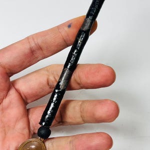 May include: A black, cylindrical amulet with silver-colored bands and a small, round, black bead. A bronze-colored oval pendant with a detailed image hangs from the amulet. The amulet is held in a hand.