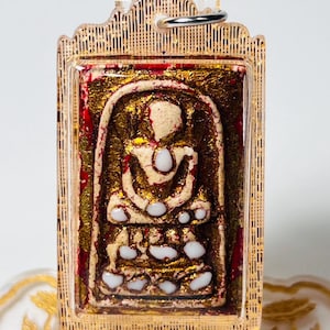 May include: A rectangular pendant featuring a seated figure with a gold and red design. The figure is in a seated position with white accents. The pendant is encased in a clear plastic frame with a gold-colored border and a silver ring at the top.