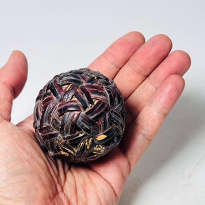 May include: A woven sphere with a dark brown and black pattern, held in an open hand. The sphere appears to be made of woven strips of material, with a slightly rough texture. The background is a plain white.
