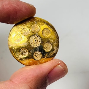 May include: A gold-colored, circular object with a series of raised, textured designs. The central design is a flower-like pattern, surrounded by smaller, circular patterns. The object is held between two fingers against a white background.